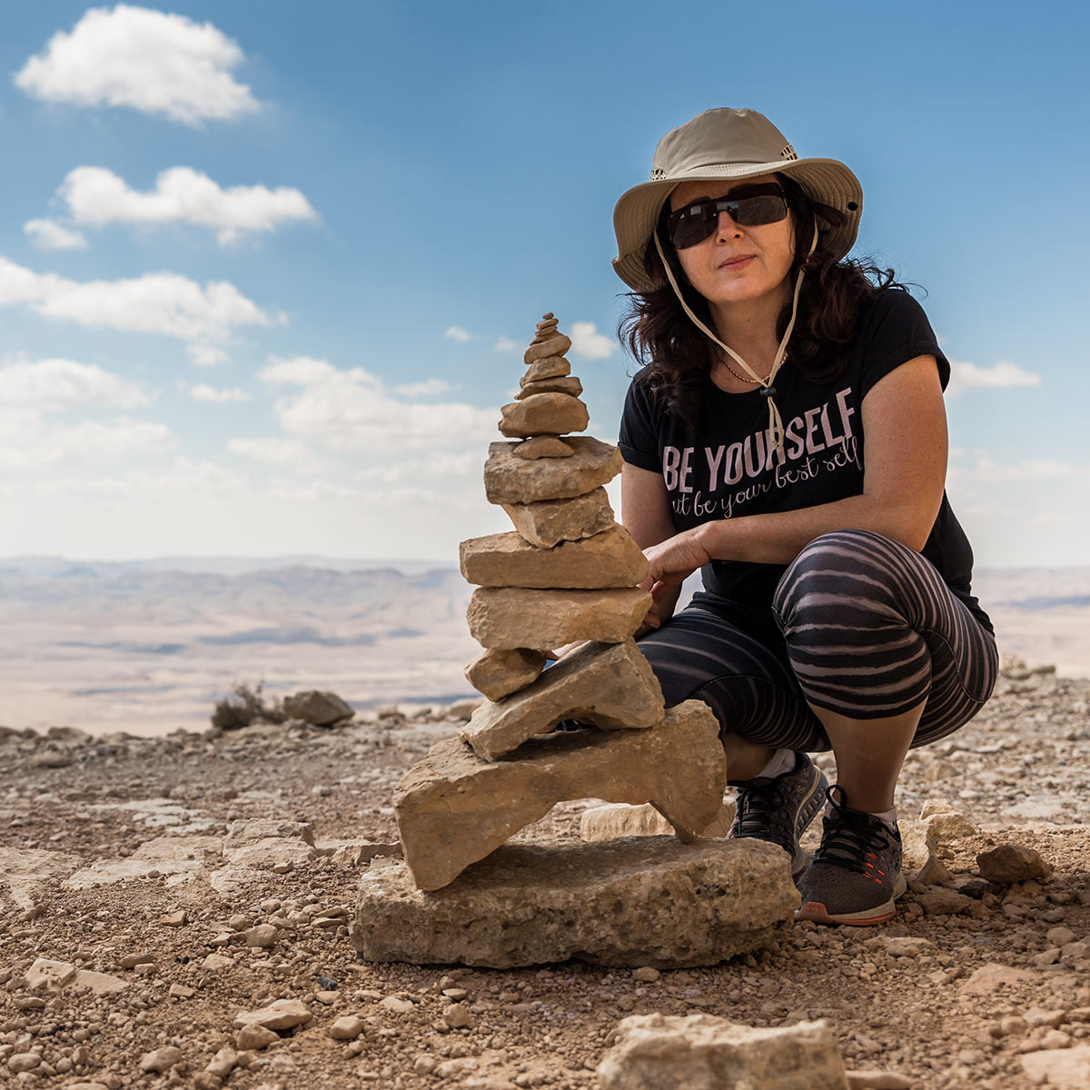 A proud woman poses in the desert sun in front of a tall rock cairn she's carefully stacked